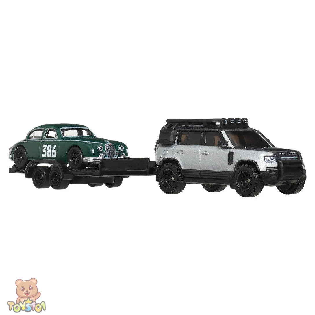 Toy Land Rover Defender with a trailer carrying a toy vintage Jaguar on a white background.